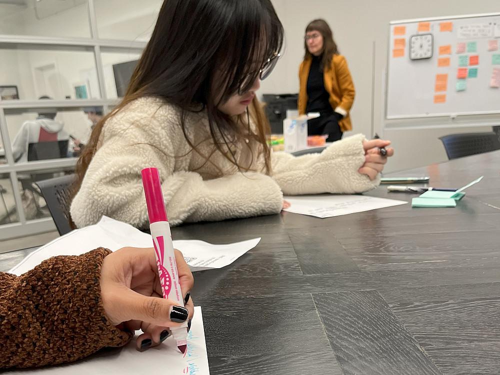 Students are sitting at a table working, while the facilitator stands behind.